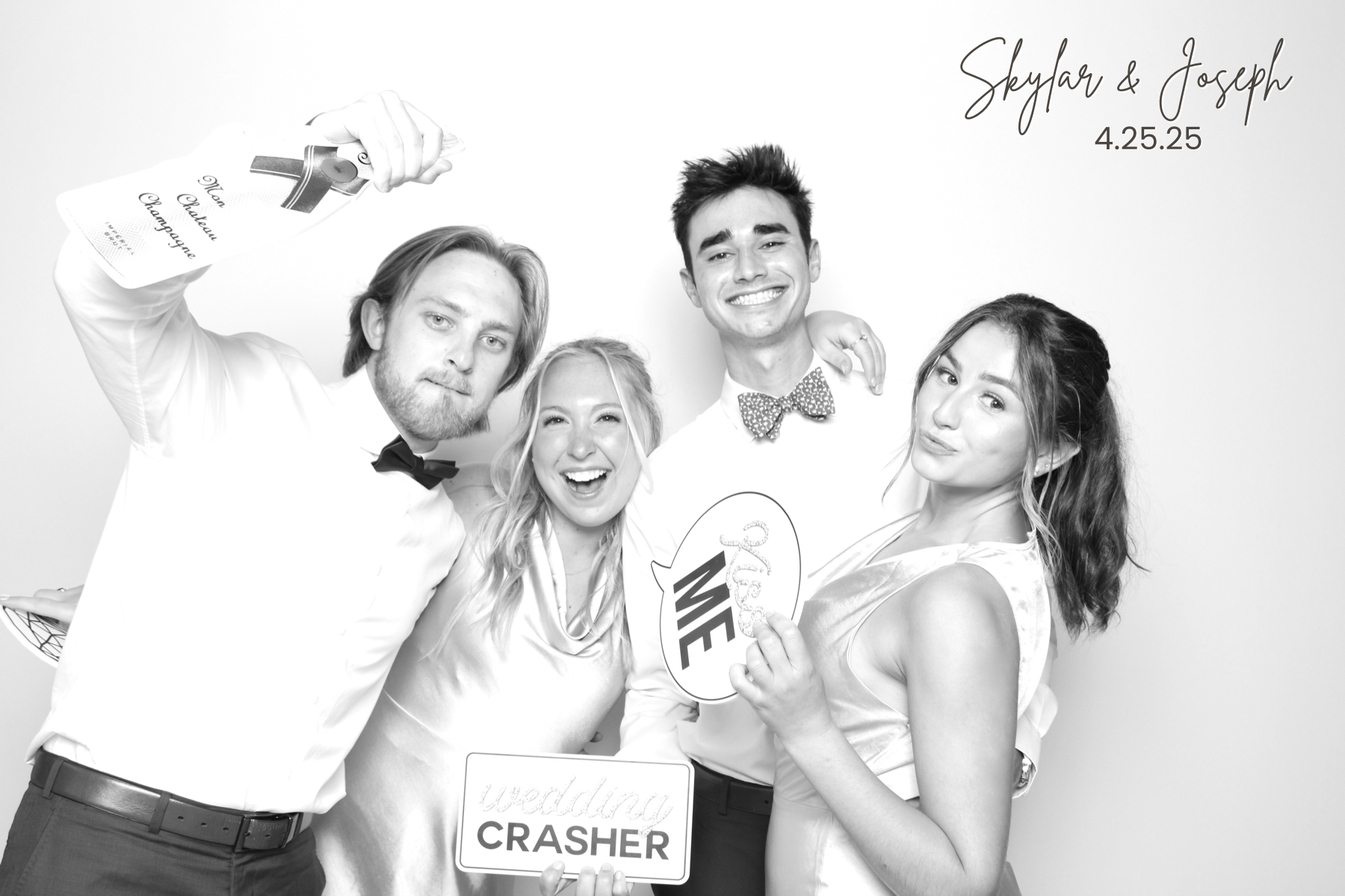 Four guests in formal attire pose with playful wedding signs and a drink.