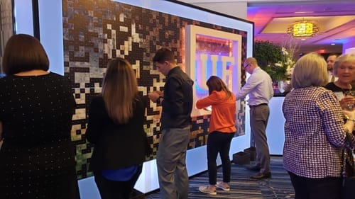 Guests placing photo tiles on a large illuminated mosaic wall at a corporate gala
