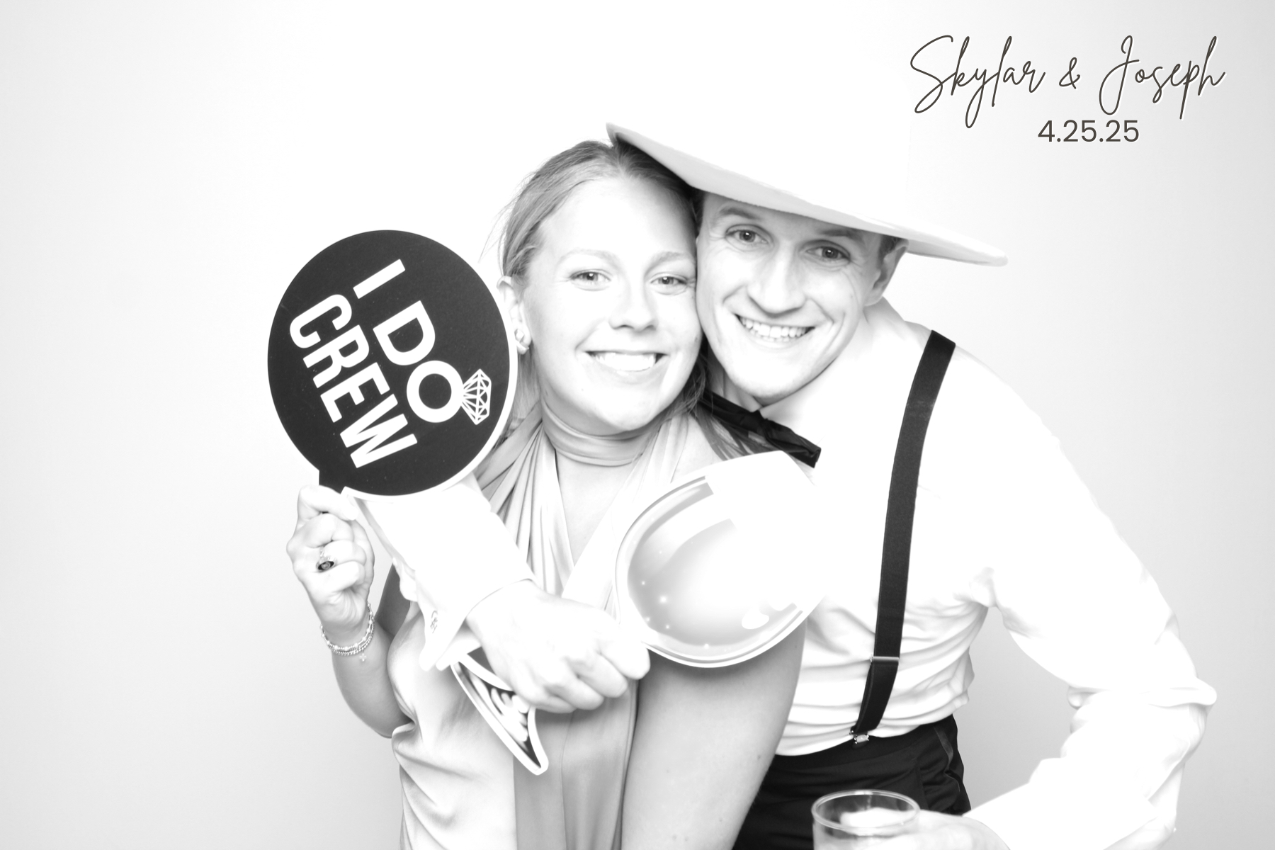 Two guests in formal attire pose closely with a champagne bottle, hat, and celebratory sign.