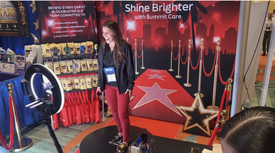 A guest laughing on the Summit Care 360 video platform in front of a Hollywood-themed "Shine Brighter with Summit Care" backdrop at the FHCA trade show in Orlando