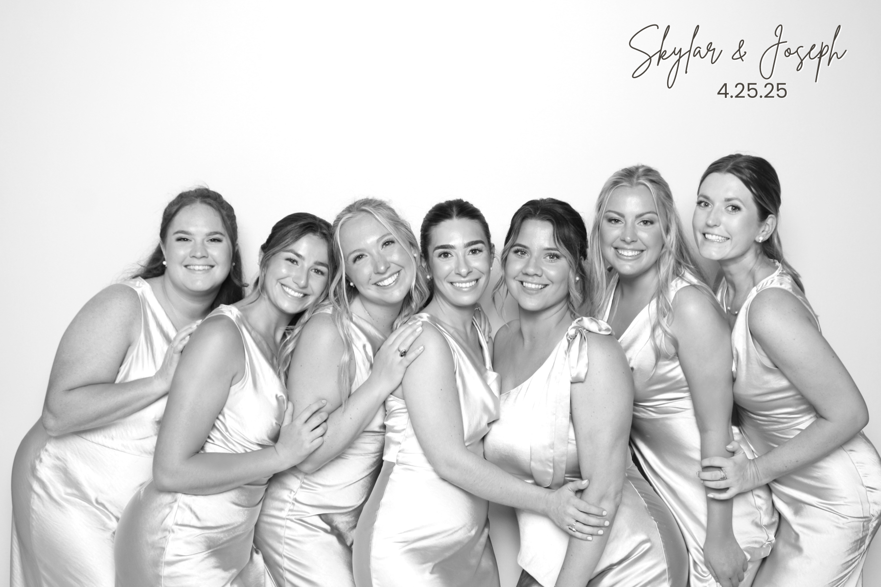 Seven wedding guests in matching satin dresses pose together in a polished black-and-white portrait.