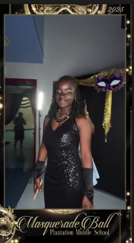 A student in a black sequin gown and lace mask posing beside the purple and gold mask display