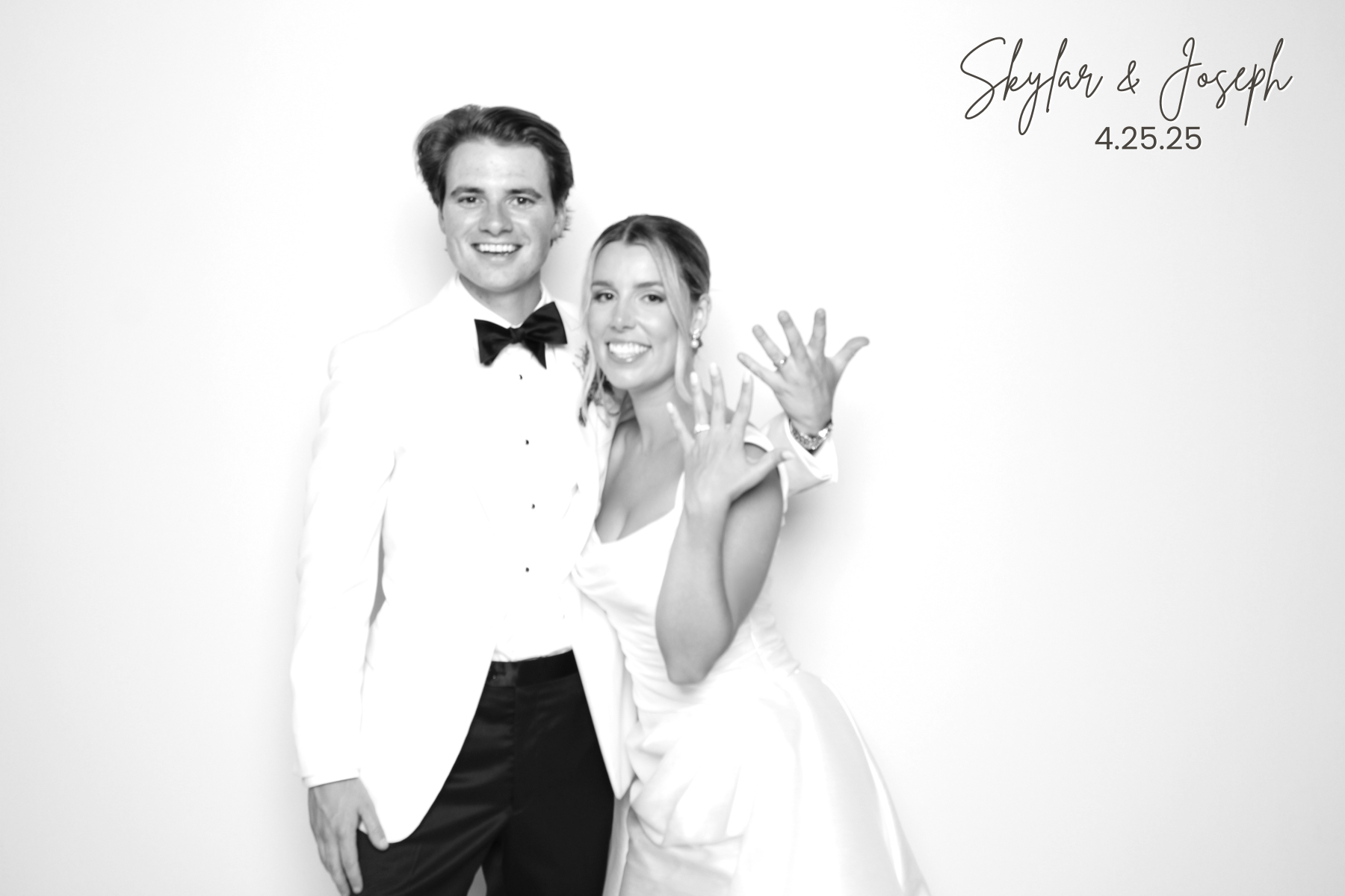 Couple in formal wedding attire smiles while holding up wedding rings against a bright studio backdrop.