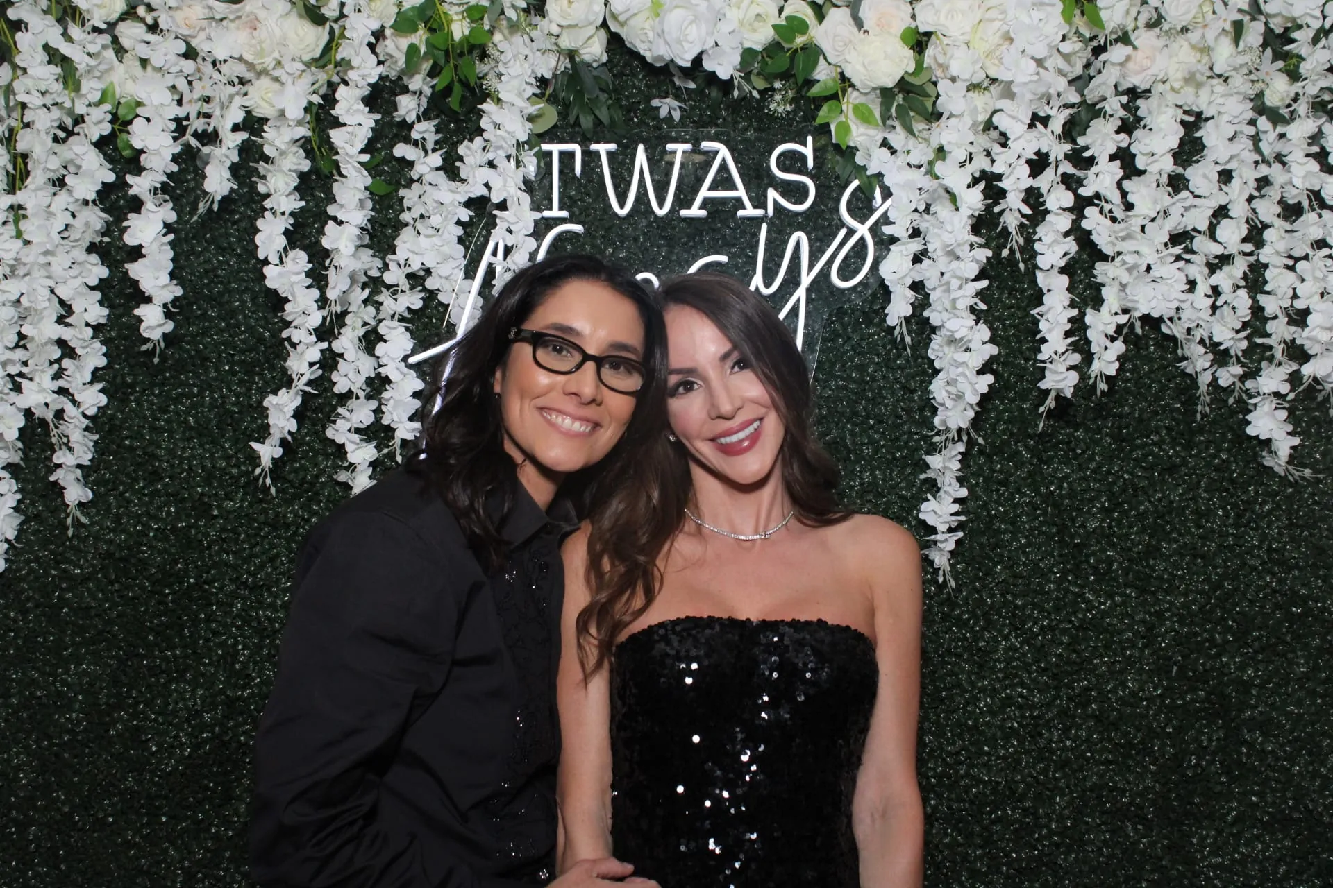 Two guests smiling in front of the greenery wall