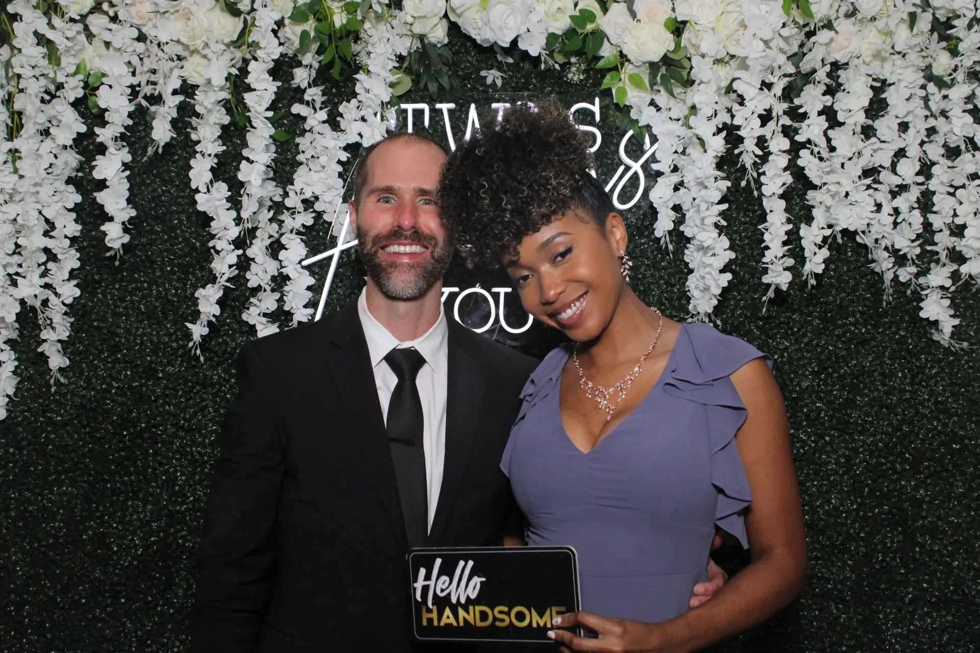 Well-dressed couple posing with a prop sign at the floral backdrop