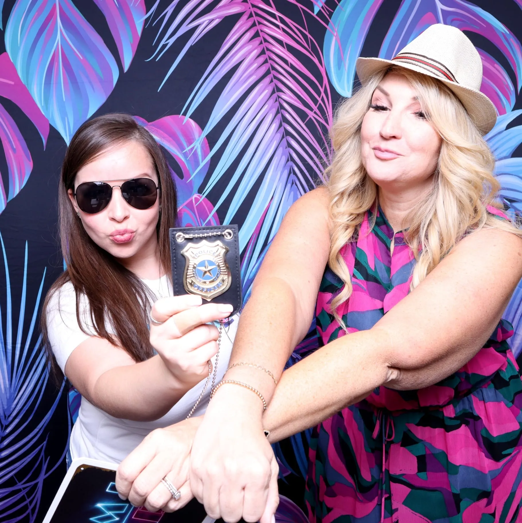 Two women with police badge prop on neon palm backdrop